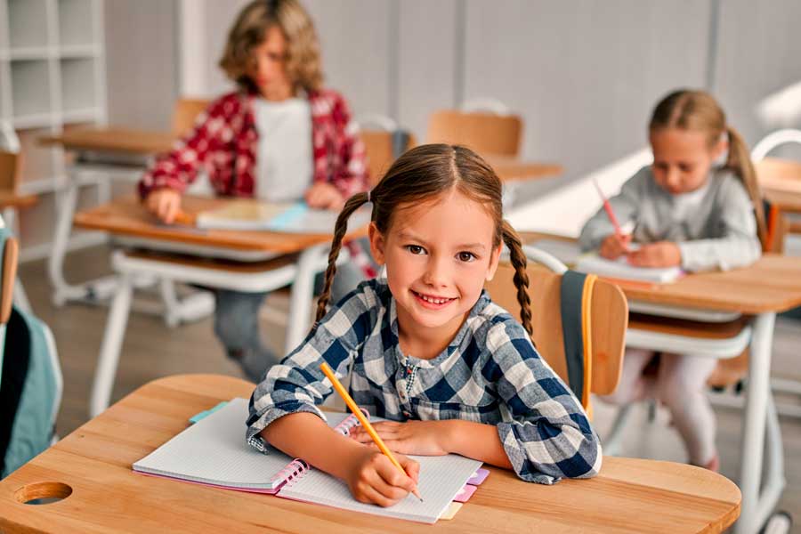 nettside_skole En smilende jente sitter ved pulten på skolen og skriver.